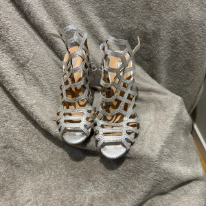 Silvriga sandaler med klack och öppna detaljer - Säljer ett par silvriga sandaler med hög klack och öppet mönster. Skorna har en snygg geometrisk design med remmar och spänne runt ankeln. Perfekta för dig som gillar att sticka ut med glittriga detaljer och elegant look.