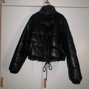 Fin jacka  - Säljer en svart pufferjacka från Glamorous i storlek 42. Jackan är croppad med snörning i midjan, har hög krage och quiltad design. Materialet är syntet med skinnlook och jackan stängs med både dragkedja och knappar. Perfekt för dig som gillar en edgy stil.