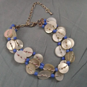 Armband med blå pärlor och knappar - Unikt armband med runda, vita knappar och blå pärlor. Tråden är tvinnad i flera färger och armbandet har en justerbar kedja i metall. Perfekt för dig som gillar boho-stil och färgglada detaljer. 19-26 cm långt.