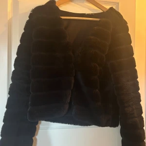 Svart pälsjacka med fluffig look Gina Tricot  - Säljer en svart pälsjacka med fluffiga, horisontella paneler och långärmad design. Jackan har en croppad passform och är tillverkad i mjukt syntetmaterial som ger en lyxig känsla. Perfekt för dig som vill ha en statement piece i garderoben. Går att knäppa.