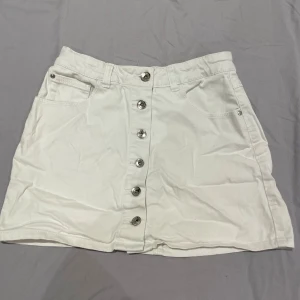 Vit kjol med knappar framtill - Snygg vit kortkjol med raka linjer och silverfärgade knappar längs hela framsidan. Kjolen har klassiska jeansfickor både fram och bak samt hällor för bälte. Perfekt för en fräsch och stilren look.