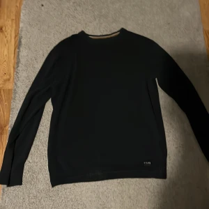 Svart långärmad tröja NYC.AVE - En stilren svart långärmad tröja från NYC.AVE med diskret logga nertill. Tröjan har rund halsringning och ribbade muddar vid ärmslut och nederkant. Perfekt för dig som gillar en grisch, enkel  och clean look.