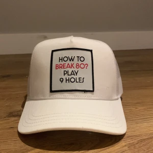 Vit keps från Bogeys & Birdies - Vit keps med svart och röd text framtill: 'How to break 80? Play 9 holes'. Kepsen har mesh-bakstycke och justerbar snapback. Tillverkad i 50% bomull och 50% polyester, perfekt för golf eller chill dagar.