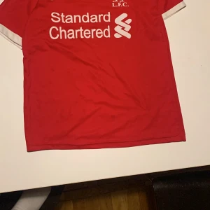 Liverpool M. Salah fotbollströja Nike - Röd Liverpool FC fotbollströja med vita detaljer och tryck från Nike. Framsidan har Standard Chartered-logga och LFC-emblem, baksidan har M. Salah och nummer 11 i vitt och gult. Klassisk rund hals och korta ärmar. Perfekt för fans av Salah och Liverpool.