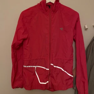 Röd vindjacka från Diadora XS - Sportig röd vindjacka från Diadora med hel dragkedja, huva och reflekterande detaljer framtill. Jackan har meshpanel på ryggen för extra ventilation och är tillverkad i lätt polyester. Perfekt för träning utomhus när det blåser.