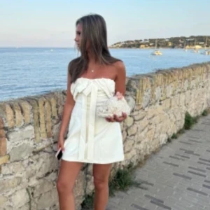 Vit axelbandslös kortklänning med rosett - Supersnygg vit axelbandslös kortklänning från Valentino med stor rosettdetalj framtill. Klänningen har en elegant och stilren design med raka linjer och är tillverkad i ett mjukt, lyxigt material som känns skönt mot huden. Klänningen är ny med prislapp!!! 