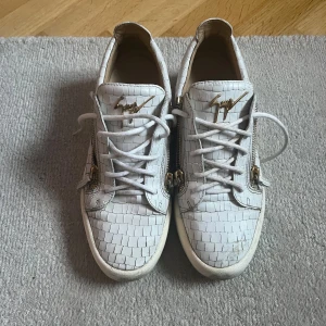 Vita sneakers från Giuseppe Zanotti - Säljer ett par vita sneakers från Giuseppe Zanotti med croco-präglat skinn, guldfärgade metalldetaljer och dragkedjor på sidorna. Skorna har snörning och rund tå, samt den ikoniska loggan i guld på plösen. Riktigt snygga och unika sneakers för dig som vill sticka ut.