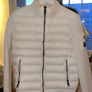 Vit Moncler pufferjacka med dragkedja - Säljer en stilren vit pufferjacka från Moncler med svarta detaljer och emblem på ärmen. Jackan har quiltad framsida, två sidofickor med dragkedja och en ficka på ärmen. Insidan har en cool serietidningsdetalj. Perfekt för dig som gillar clean och exklusiv look.