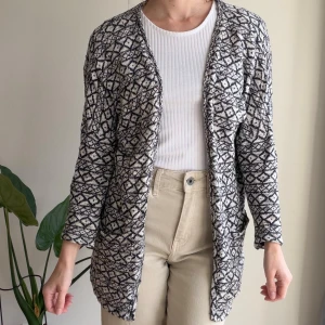 Mönstrad kofta från Pieces - Säljer en blåvit stickad kofta från Pieces med grafiskt mönster. Koftan har lång ärm, öppen front och två fickor framtill. Perfekt att slänga över en enkel topp för en chill och snygg look.