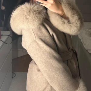 Beige kappa med pälsdetaljer  - Säljer en beige kappa från femenina med fluffiga pälsdetaljer på krage och ärmslut. Jackan har omlottmodell med bälte i midjan och en guldfärgad logga på ärmen. Perfekt för dig som vill ha en lyxig och varm look i vinter. Alla pälsdetaljer är avtagbara