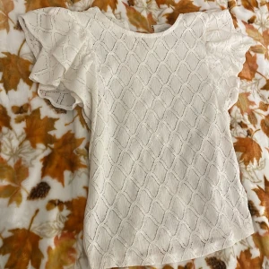 Vit blus med volangärm Holly & Whyte - Supersöt vit blus från Holly & Whyte med strukturmönster och volangärmar. Blusen har rund halsringning och är tillverkad i ett lätt, luftigt tyg som känns fräscht och somrigt. Perfekt för dig som gillar romantiska detaljer och en feminin look.