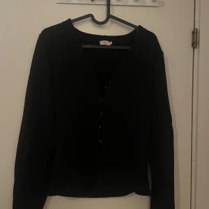 Svart kofta från Nelly med knappar - Svart tröja från Nelly i storlek M. Den har en enkel och clean design med v-ringad hals och knappar framtill. Långärmad och passar perfekt när du vill ha en stilren look. Materialet känns mjukt och skönt mot huden.