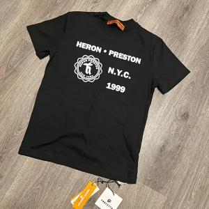 Svart Heron Preston t-shirt NYC 1999 - Svart t-shirt från Heron Preston med vit NYC 1999-tryck och logga på framsidan. Baksidan har en stor färgglad grafisk print med fåglar och stjärnor. Klassisk rund hals och korta ärmar. Materialet är mjuk bomull och har orange etikett i nacken.