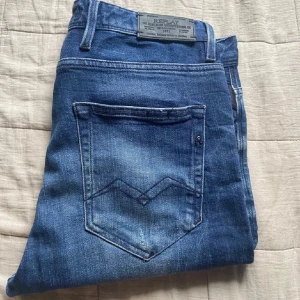Replay Anbass 29/32 - Säljer ett par Replay Anbass jeans i blå tvätt med snygga slitningar och klassiska bakfickor med V-detalj i storlek 29/32. Jeansen har skinny passform och är tillverkade i stretchigt jeanstyg för extra komfort. Perfekta för dig som gillar en smal siluett och stilren look.
