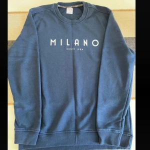 Marinblå sweatshirt från MLN Milano - Snygg marinblå sweatshirt från MLN Milano med vit text 'MILANO SINCE 1984' på bröstet. Tröjan har rund hals, ribbade muddar och lång ärm. Perfekt för dig som gillar enkel och stilren design med en urban touch. Köpt i Italien.