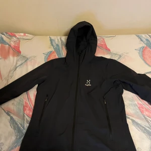 Svart vindjacka från Haglöfs med huva - Svart vindjacka från Haglöfs med stilren design och praktisk huva. Jackan har två dragkedjeförsedda fickor på framsidan och diskret Haglöfs-logga på bröstet. Långärmad och tillverkad i slitstark syntet, perfekt för blåsiga dagar.