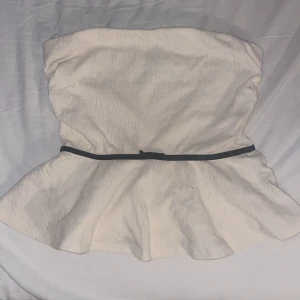 Krämvit tubtopp med svart rosett - Säljer en krämvit tubtopp från Holly & Whyte by Lindex i storlek M. Toppen är axelbandslös och har en svart smal bälte med en liten rosett i midjan. Materialet är mjukt och har en lätt struktur som ger en cool känsla. Perfekt för dig som gillar stilrena detaljer.