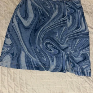 Blå swirlmönstrad kortkjol  - Säljer en blå kortkjol med unikt swirlmönster i olika blå nyanser. Kjolen har en liten slits framtill och dragkedja bak. Sitter snyggt och har en tight passform som framhäver midjan. Från en amerikansk affär ”target” 