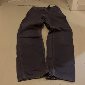 Mörkgrå cargobyxor från Carhartt - Säljer ett par mörkgrå cargobyxor från Carhartt med bred passform och flera praktiska fickor på sidorna och bak. Byxorna har klassisk Carhartt-logga på bakfickan och är tillverkade i slitstark bomull. Perfekta för dig som gillar streetwear och funktionella plagg.