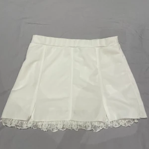 Vit kortkjol med spetskant - Supersöt vit kortkjol med rak passform och dekorativ spetskant längst ner. Kjolen har en enkel design med diskreta sömmar framtill och är perfekt för dig som gillar en clean och feminin stil.