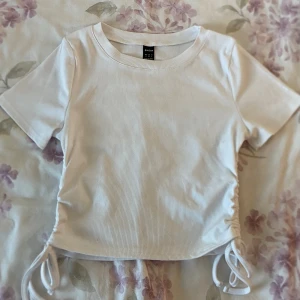 Ribbad croppad topp - Säljer en vit ribbad croppad topp från SHEIN i storlek S. Toppen har korta ärmar och snygga snörningsdetaljer på båda sidorna som går att justera för en cool look. Perfekt för dig som gillar stilrena och trendiga plagg.