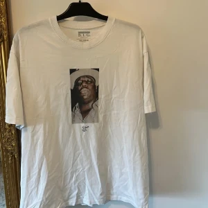 Vit Notorious B.I.G. t-shirt Pull&Bear - Vit t-shirt från Pull&Bear med tryck av Notorious B.I.G. på bröstet. Klassisk rund halsringning och korta ärmar. Perfekt för dig som gillar streetwear och hiphop-inspirerad stil.