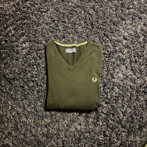 Svart v-ringad tröja Fred Perry - Svart v-ringad tröja från Fred Perry i storlek L. Klassisk design med ribbade muddar och en diskret Fred Perry-logga broderad på bröstet. Tröjan har en enkel och stilren look med en tunn, mjuk känsla som passar perfekt till jeans eller chinos.