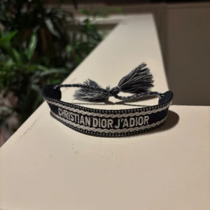Christian Dior J'Adior armband - Snyggt armband från Christian Dior med texten 'J'Adior' broderad i vitt på svart bakgrund. Armbandet har justerbar knytning med tofsar i ändarna och en cool, vävd design som ger en exklusiv känsla.