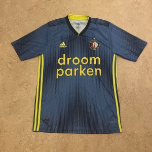 Feyenoord 19/20 bortatröja Adidas M - Feyenoord bortatröja från säsongen 19/20 i marinblått med gula detaljer och prickigt mönster. Adidas-logga och klubbmärke på bröstet, samt sponsortryck 'droom parken' framtill. Tillverkad i lätt och ventilerande funktionsmaterial. I storlek M ete kvalité 