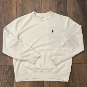 Vit sweatshirt från Polo Ralph Lauren - Vit sweatshirt från Polo Ralph Lauren med klassisk rund hals och broderad logga på bröstet. Tröjan har långa ärmar och ribbade muddar vid ärmslut och nederkant. Perfekt för en clean och stilren look.