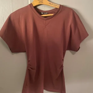 Brun kort klänning från Zara - Kort klänning från Zara i en snygg brun nyans. Modellen har korta ärmar, rund halsringning och lätt rynkade detaljer i midjan för en cool siluett. Tillverkad i mjuk bomull som känns skön mot huden. Perfekt för dig som gillar enkel och stilren design.