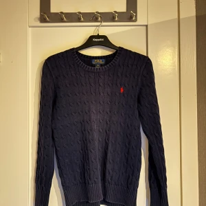 Marinblå stickad tröja Polo Ralph Lauren - Snygg marinblå stickad tröja från Polo Ralph Lauren med klassiskt kabelstickat mönster och röd broderad logga på bröstet. Rund halsringning och långa ärmar, perfekt för dig som gillar stilrena och tidlösa plagg. Storlek L (14-16) men sitter som en vuxen XS-S. 