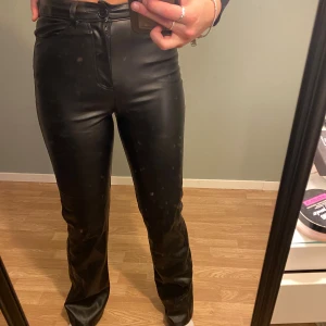 Svarta utsvängda byxor i skinnimitation - Säljer ett par svarta byxor i skinnimitation med lite utsvängdning och hög midja. Byxorna har klassiska fickor fram och bak, bälteshällor och stängs med dragkedja och knapp. Snyggt glansigt material som ger en cool look.
