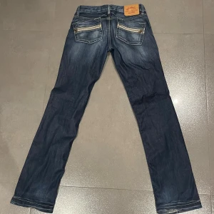  jeans med dragkedjor  - Snygga blå jeans med raka ben och coola dragkedjor på bakfickorna. Jeansen har klassisk femficksdesign, kontrastsömmar och en läderpatch bak i midjan. Perfekta för dig som gillar detaljer och vill sticka ut lite extra.