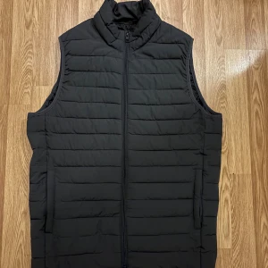 Svart/grå dunväst från Jack & Jones - Stilren svart dunväst från Jack & Jones med quiltad design. Västen har dragkedja framtill och två sidofickor. Tillverkad i syntetmaterial. Knappt använd. Hör gärna av er vid köp och frågor.