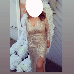 Glittrig guldig långklänning med slits - Säljer en superglittrig långklänning i guld med v-ringning, långa ärmar och en snygg slits framtill. Klänningen har figurnära passform och är täckt av paljetter som verkligen fångar ljuset. Perfekt för dig som vill sticka ut på bal eller fest! kan diskutera pris!