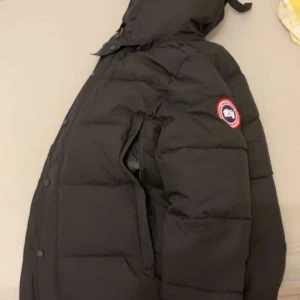 Svart dunjacka från Canada Goose - Svart dunjacka från Canada Goose med huva och avtagbar pälsdetalj. Jackan har klassisk patch på ärmen, dragkedja och knappar framtill. Fylld med dun och har en loose passform, perfekt för kalla vinterdagar. Snygg och tidlös design som håller dig varm. Storlek M 