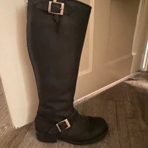 Johnny bulls  - Snygga svarta höga boots med två silvriga spännen och grov dragkedja baktill. Klassisk rund tå och låg klack, perfekt till jeans eller kjol. Bootsens stilrena design gör dem lätta att matcha och de har en robust känsla.