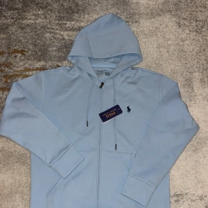 Ljusblå hoodie från Polo Ralph Lauren - Snygg ljusblå hoodie från Polo Ralph Lauren med dragkedja framtill och klassisk huva. Tröjan har justerbara snören vid huvan och den ikoniska broderade loggan på bröstet. Perfekt för en chill och stilren look.