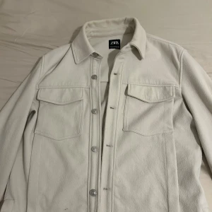 Beige overshirt från Zara - Stilren beige overshirt från Zara med klassisk krage, två stora bröstfickor och silverfärgade tryckknappar. Skjortan är i ett mjukt material som känns lite fleeceaktigt och har lång ärm. Perfekt för lager-på-lager och enkel att matcha.