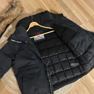  Zavetti Canada puffar jacka  - Svart pufferjacka från Zavetti Canada med avtagbar fuskpäls på huvan, quiltad insida och flera fickor framtill. Jackan har ribbade muddar, dragkedja och snygga logodetaljer på axel och insida. Perfekt för kalla dagar och har en modern, clean look.