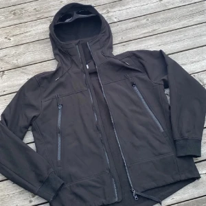 Svart Goggle Jacket från C.P. Company - Svart Goggle Jacket från C.P. Company med ikoniska glasögon i huvan. Jackan har två stora dragkedjefickor fram, lång ärm och en clean, sportig look. Materialet är mjukt och syntetiskt, perfekt för dig som gillar streetwear och vill sticka ut.