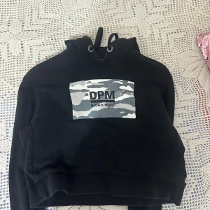 Svart hoodie från Björn Borg, croppad - Svart croppad hoodie från Björn Borg med huva och snörning. Framsidan har ett grått och vitt camouflage-tryck med texten DPM. Tröjan har långa ärmar och är tillverkad i mjuk bomullsmix, perfekt för chill eller streetwear.