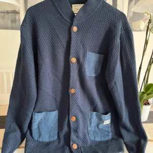 Mörkblå kofta från Jack & Jones XL - Snygg mörkblå kofta från Jack & Jones i storlek XL. Koftan har ett coolt strukturerat stickat tyg, tre blå manchesterfickor och träknappar framtill. Ribbad krage och muddar ger extra stilpoäng. Perfekt för dig som gillar detaljer och chill vibe.