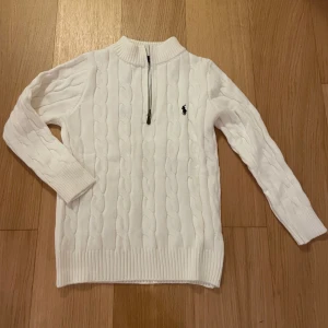 Vit stickad 1/4 zip Polo Ralph Lauren - Säljer en vit stickad tröja från Polo Ralph Lauren med kabelstickat mönster och halv dragkedja vid halsen. Tröjan har långärmad design, ribbade muddar och klassisk broderad logga på bröstet. Perfekt för dig som gillar stilren och tidlös look.