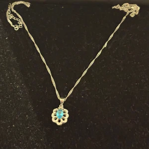Äkta Silverhalsband med blå sten - Ett elegantÄkta silverhalsband med tvinnad kedja och ett hänge i form av en blomma. Hänget har en klar blå sten i mitten, omgiven av små detaljer i silver. Märkt 925 för äkta silver. Perfekt för dig som gillar klassiska smycken med färgklick.