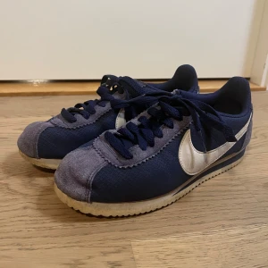 Nike Cortez i marinblå och vitt - Säljer ett par klassiska Nike Cortez sneakers i marinblått med vita detaljer och sula. Ovandelen är i textil med mockadetaljer på tå och sidor, samt den ikoniska vita swoosh-loggan. Skorna har snörning och platt sula för en retro vibe. Sulorna behöver limmas om. 