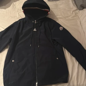 Mörkblå vindjacka från Moncler - Snygg mörkblå vindjacka från Moncler med huva och coola detaljer som snörning och dragkedja framtill. Jackan har Moncler-logga på ärmen och silverfärgade knappar vid fickorna. Perfekt för dig som gillar stilren design och exklusiva märken. Lappar till jackan finns också!
