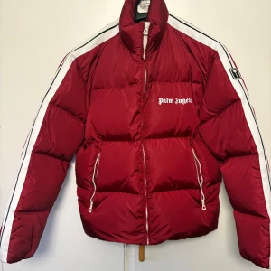 Röd Palm Angels pufferjacka - Säljer en fet röd pufferjacka från Palm Angels med vita och svarta stripes längs ärmarna, hög krage och logga på bröstet. Jackan har dragkedja framtill och på fickorna, samt innerficka. Materialet är polyester och dun, perfekt för kalla dagar.