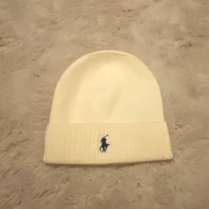 Vit stickad mössa från Polo Ralph Lauren - Vit, ribbstickad mössa från Polo Ralph Lauren med klassisk broderad logga framtill. Enkel och clean design med uppvikt kant, perfekt för kalla dagar. Mjuk och skön känsla som passar till alla dina outfits.